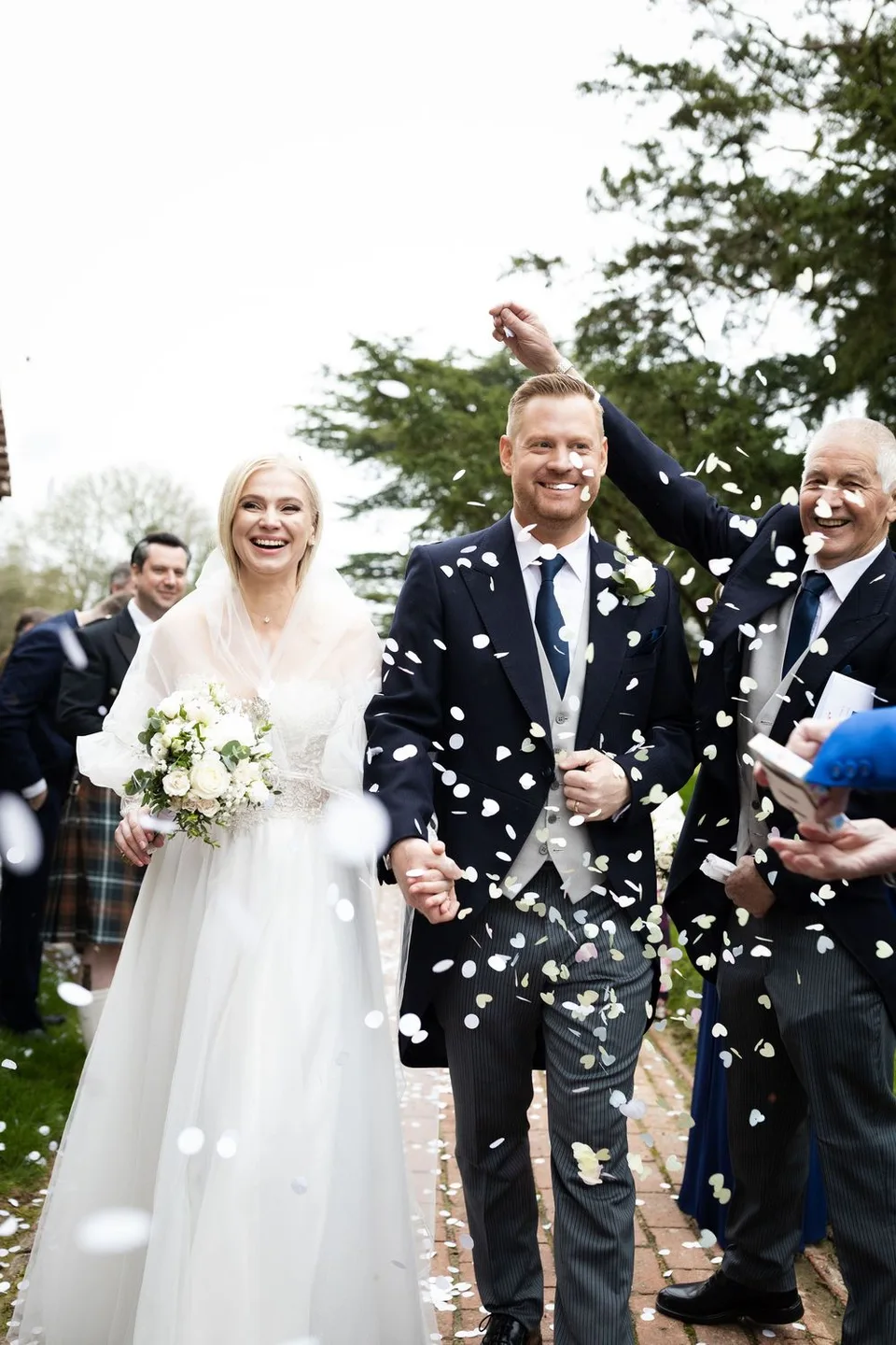 Joyful newlyweds walk through confetti shower, bride in white gown holding bouquet, groom in dark suit, guests celebrating outdoors.