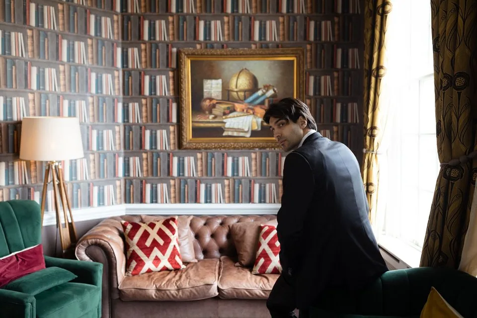Person in dark suit seated on leather couch in library with bookshelf wallpaper.