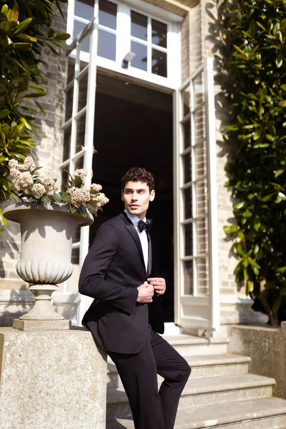 Elegant formal portrait of a groom in black tuxedo standing by stone stairs and decorative urn planter.