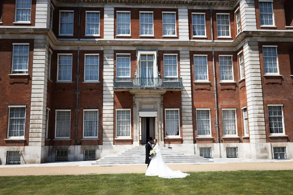 Bride and groom pose before a grand red brick Georgian manor house with white stone columns on a sunny day.