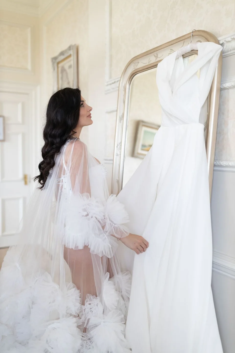 Person in sheer robe admiring white wedding dress hanging on vintage mirror frame.