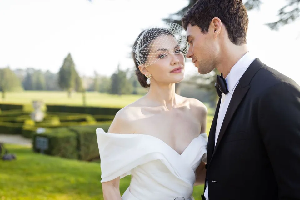 Elegant wedding couple poses in formal attire against a sunlit garden backdrop.