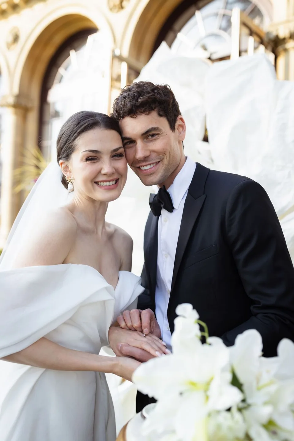 A wedding couple shares an intimate moment against a backdrop of classic architecture and white flowers.