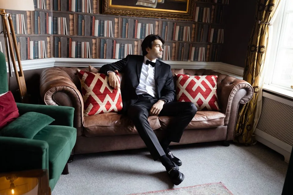 Person in tuxedo relaxes on vintage leather couch in library with book-lined walls.