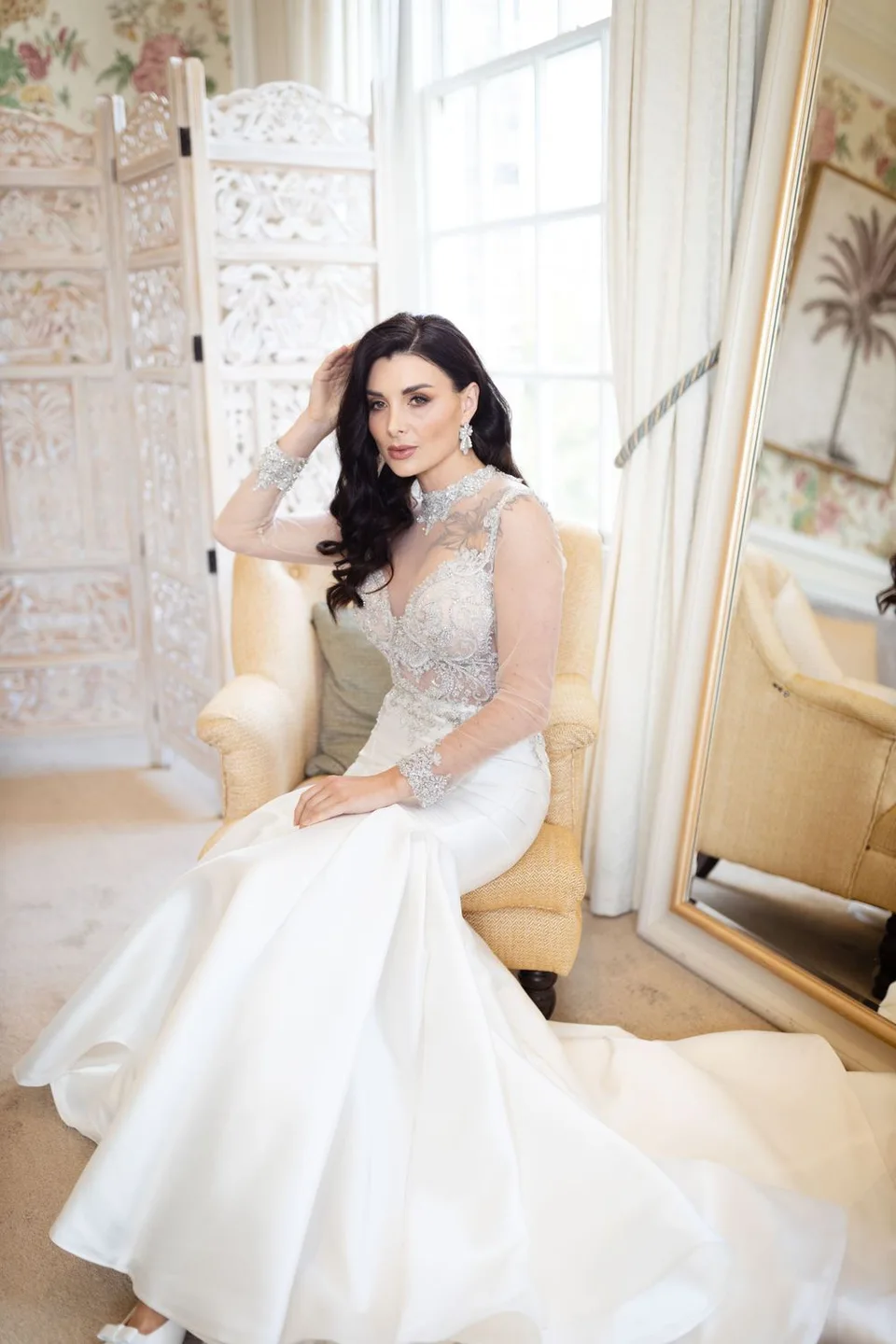 A stunning white lace wedding dress displayed in an elegant room with floral wallpaper and ornate decor.