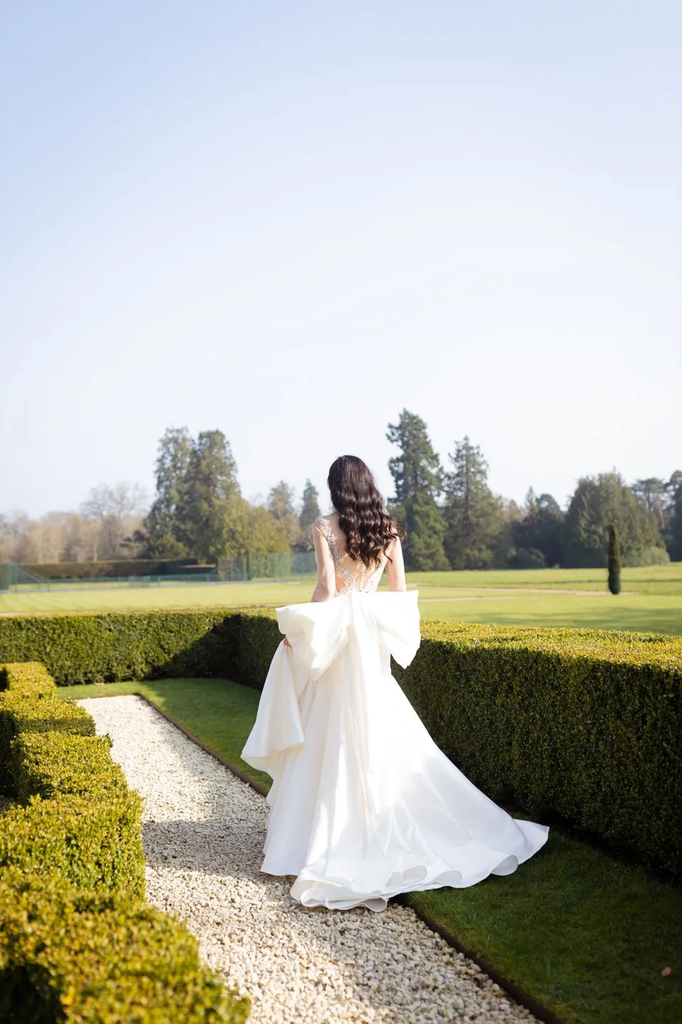 Bride in flowing white dress walks along manicured garden path during golden hour.