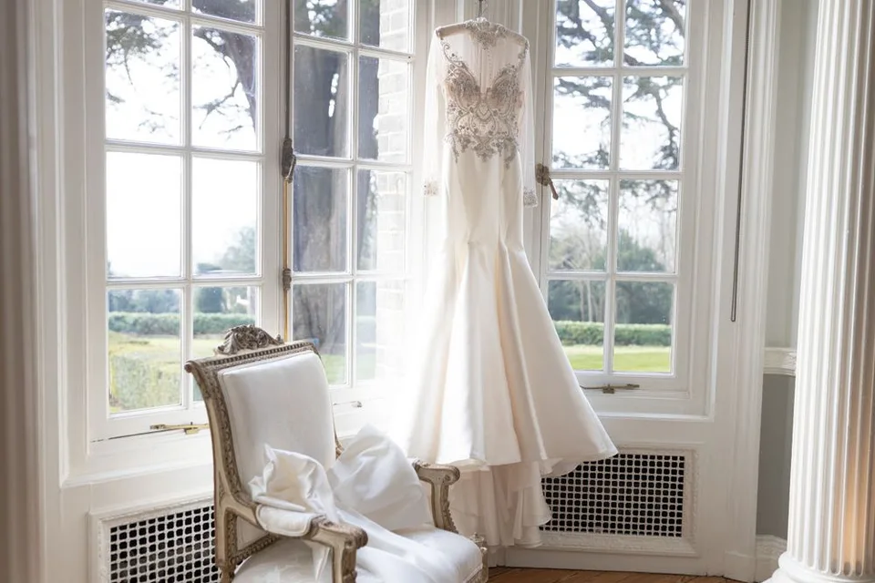 Elegant white wedding dress hanging by window with lace details and long train.