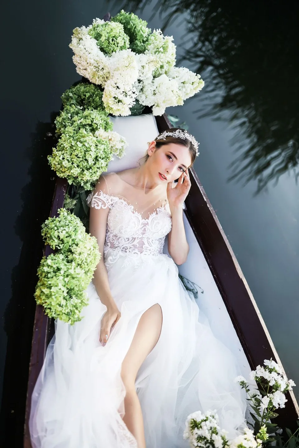 Elegant white wedding gown adorned with green and white hydrangea flowers in a wooden boat on calm water.
