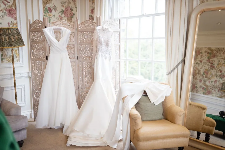 White wedding gowns displayed on hangers in an elegant dressing room with floral wallpaper.