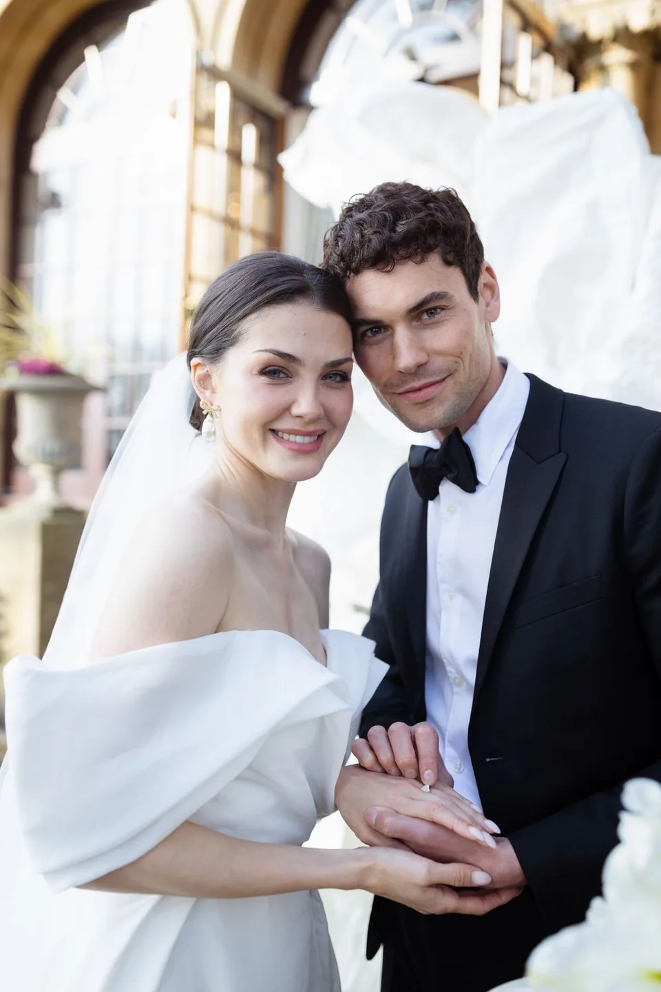 A bride in an elegant off-shoulder gown poses with her groom in front of an ornate architectural backdrop.