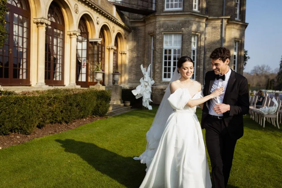 Newlyweds share a moment outside a grand stone mansion with arched windows and manicured gardens.