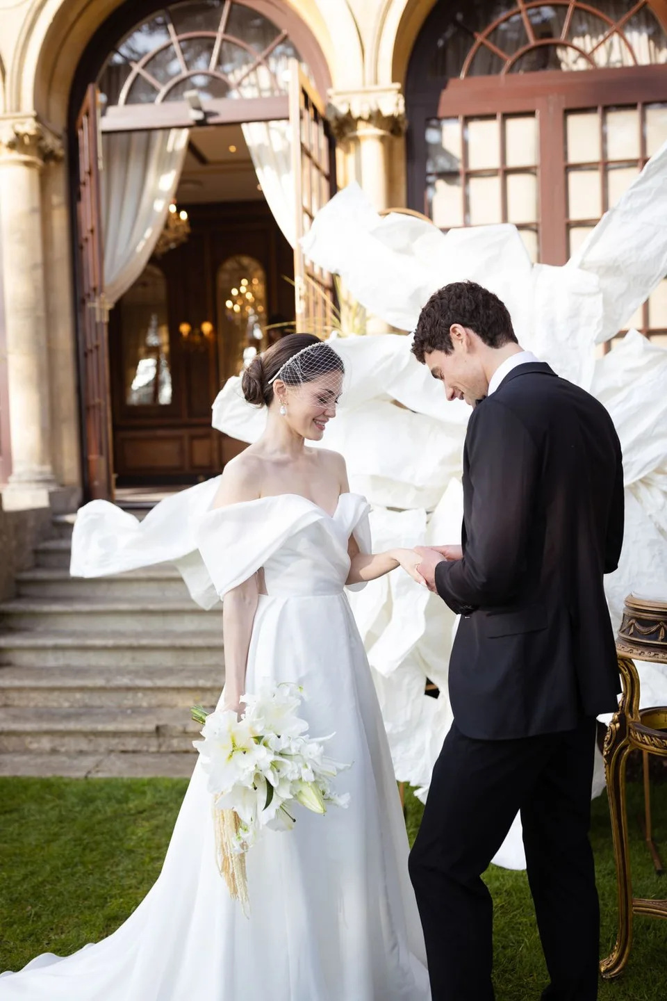 A wedding couple shares a moment on the lawn in front of a grand historic building with arched windows.