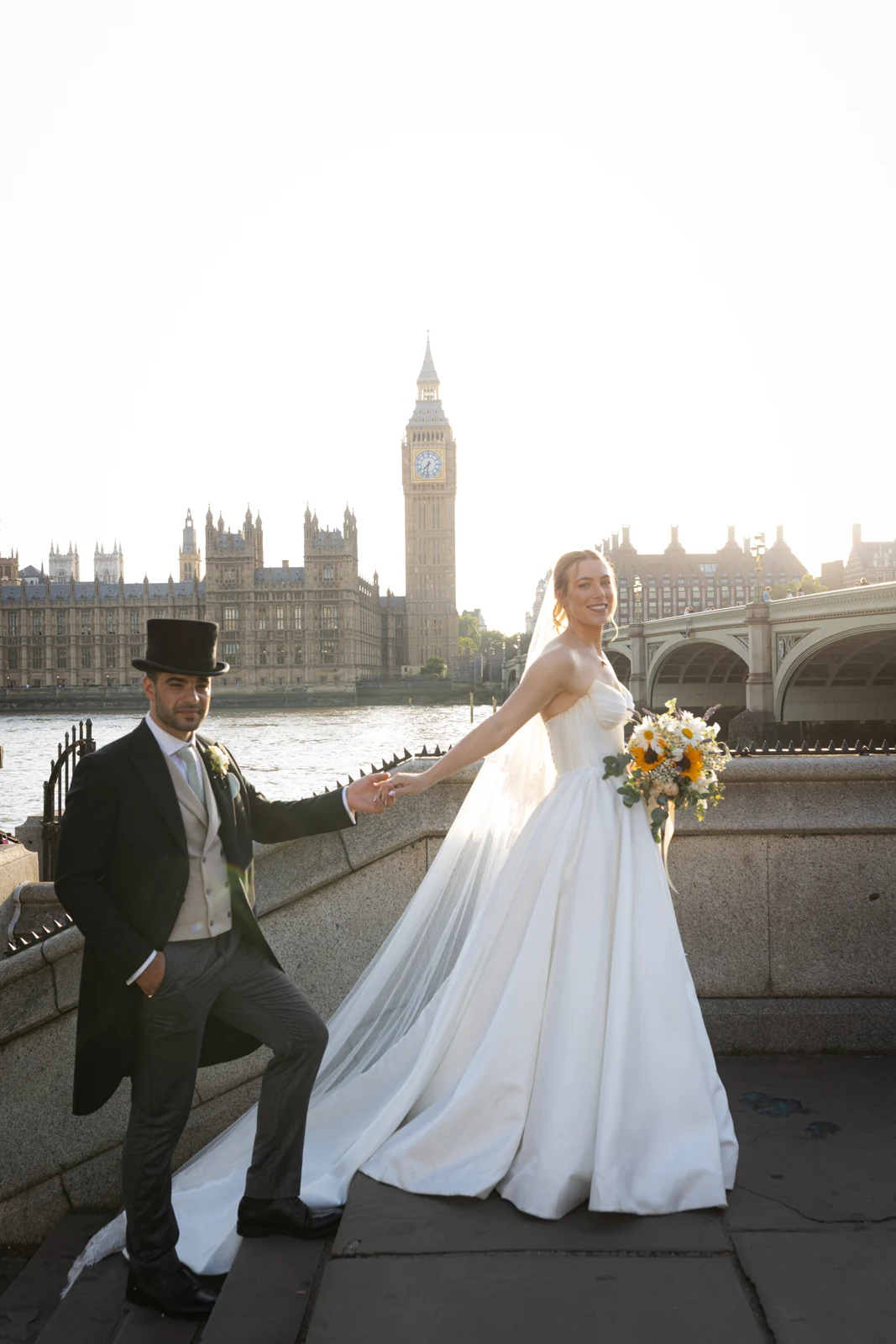 A wedding couple holding hands beside the Thames, with Westminster Bridge and Big Ben behind them.
