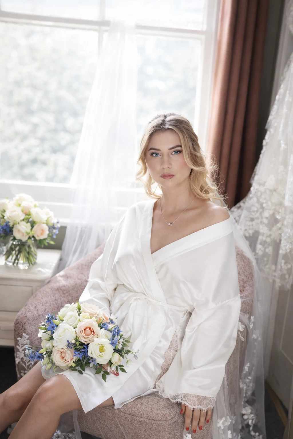 A bride in a white robe seated beside a window with her bouquet.