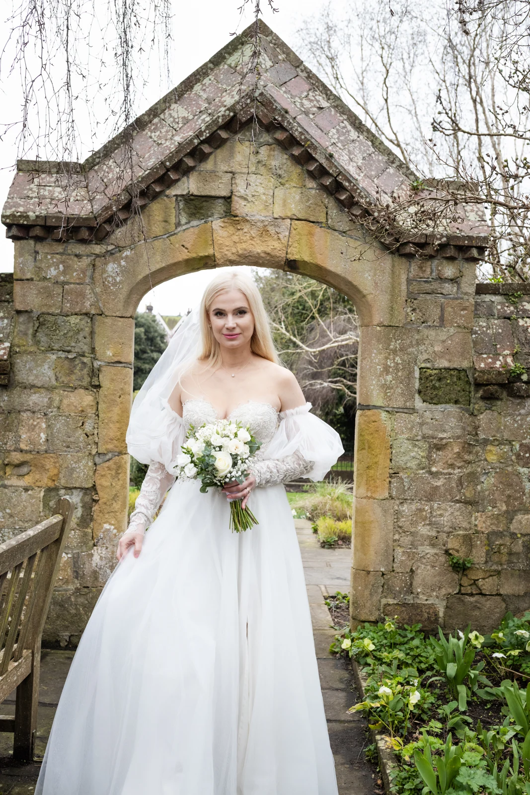 A bride holding a white bouquet beneath a stone garden archway.