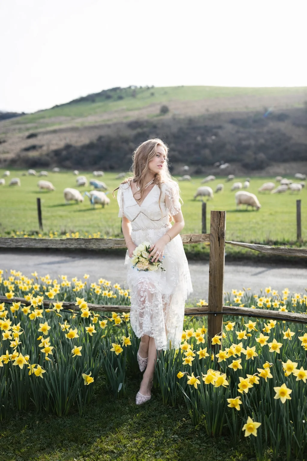 Bride holding a bouquet among daffodils, with sheep grazing on the hillside behind her