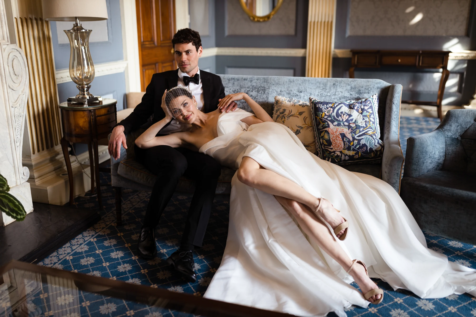 Bride reclining beside the groom on a blue sofa in an elegant room.