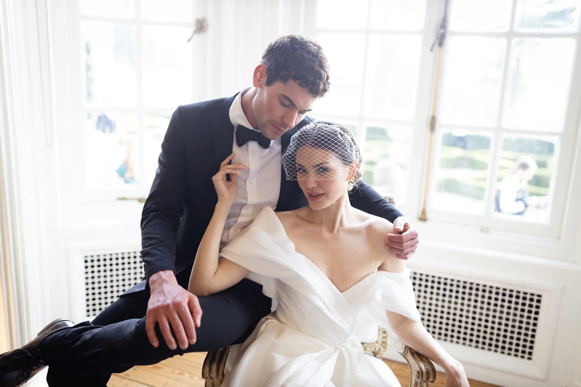 Bride and groom seated closely together beside a bright window.