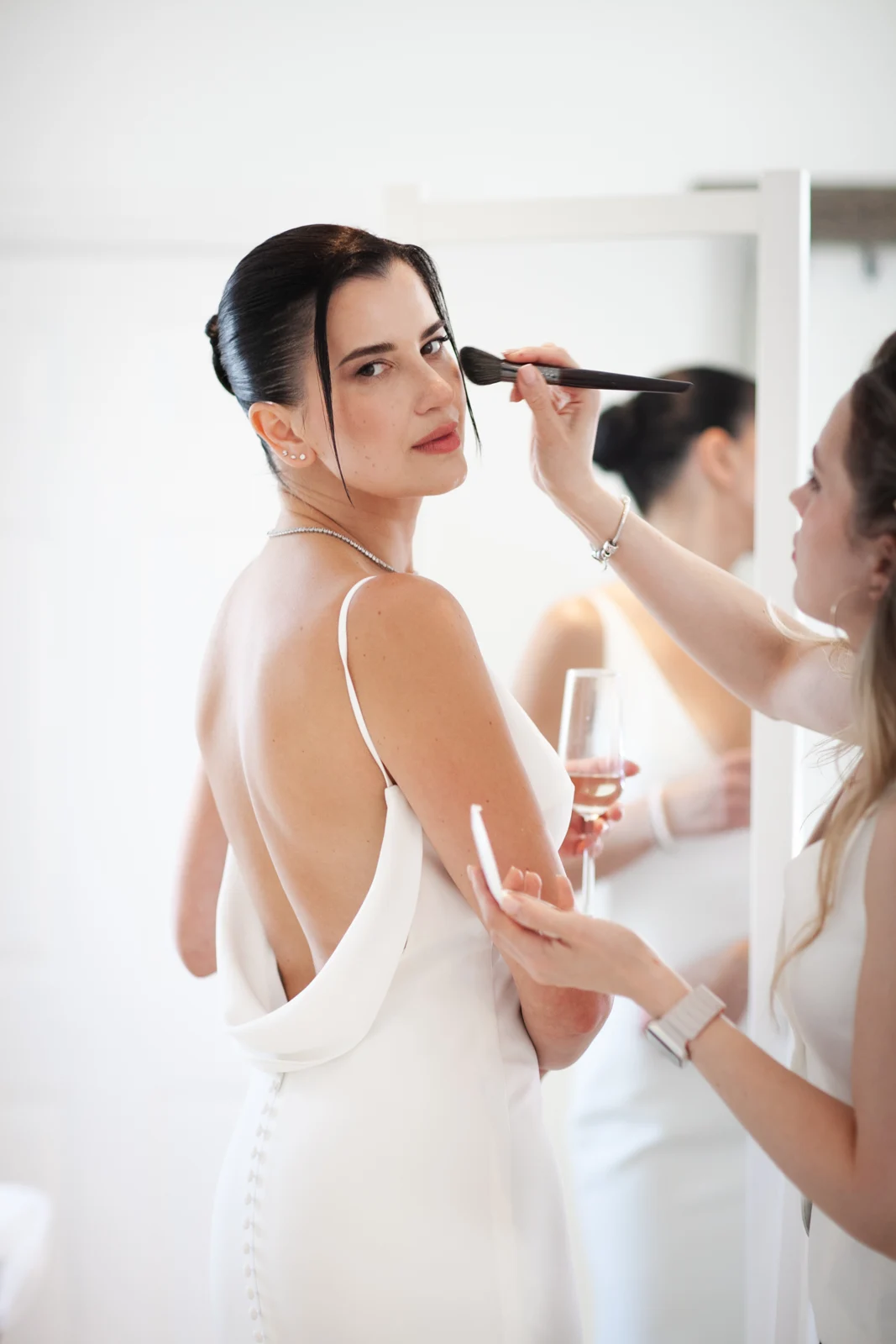 A bride holding a glass while a makeup artist applies the finishing touches.