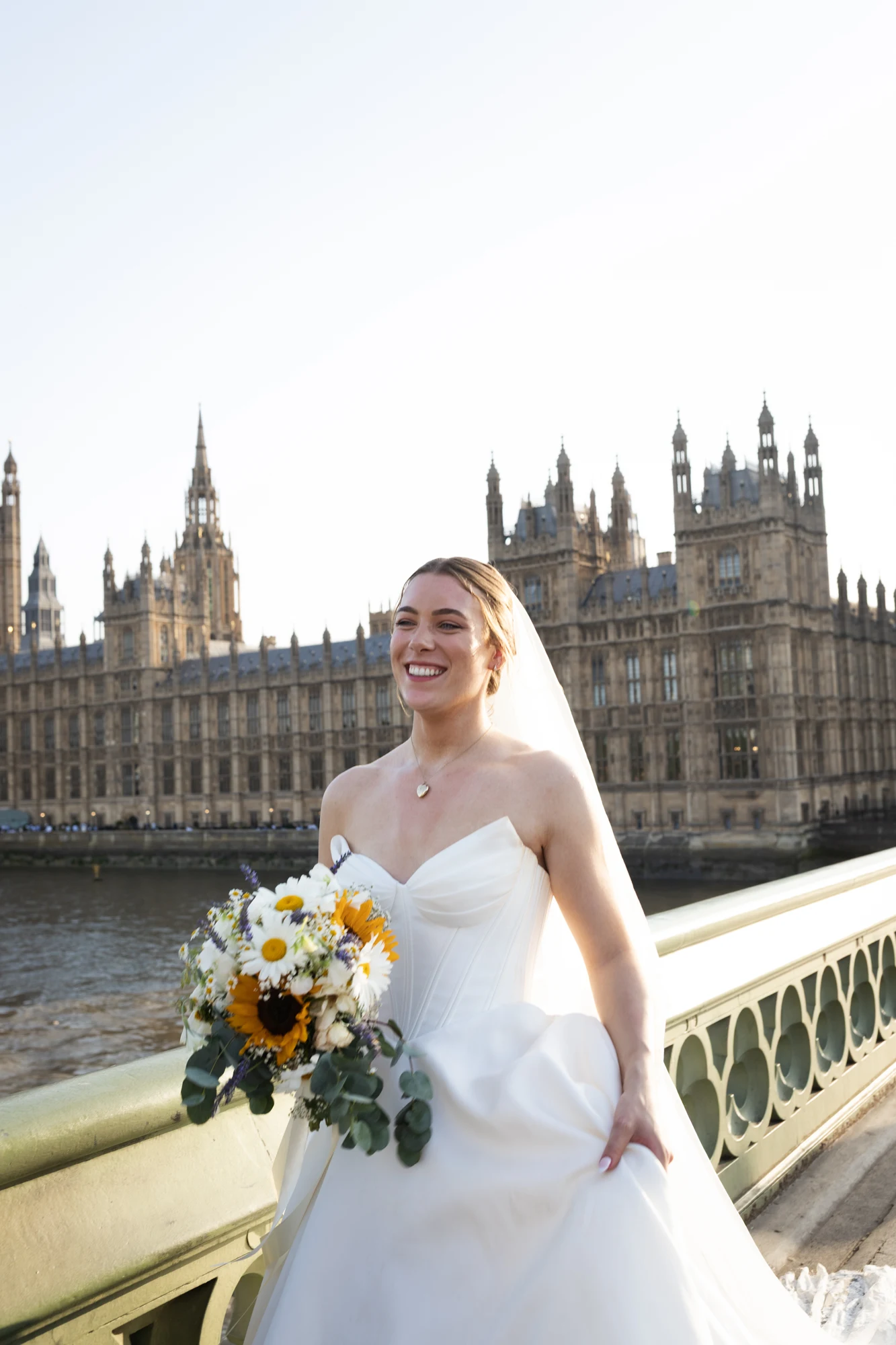 Smiling bride carrying a sunflower bouquet on Westminster Bridge in warm evening light.