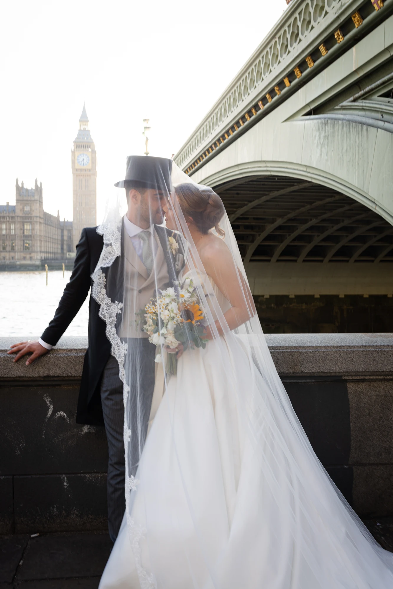 Bride and groom sharing a kiss beneath her veil beside Westminster Bridge.