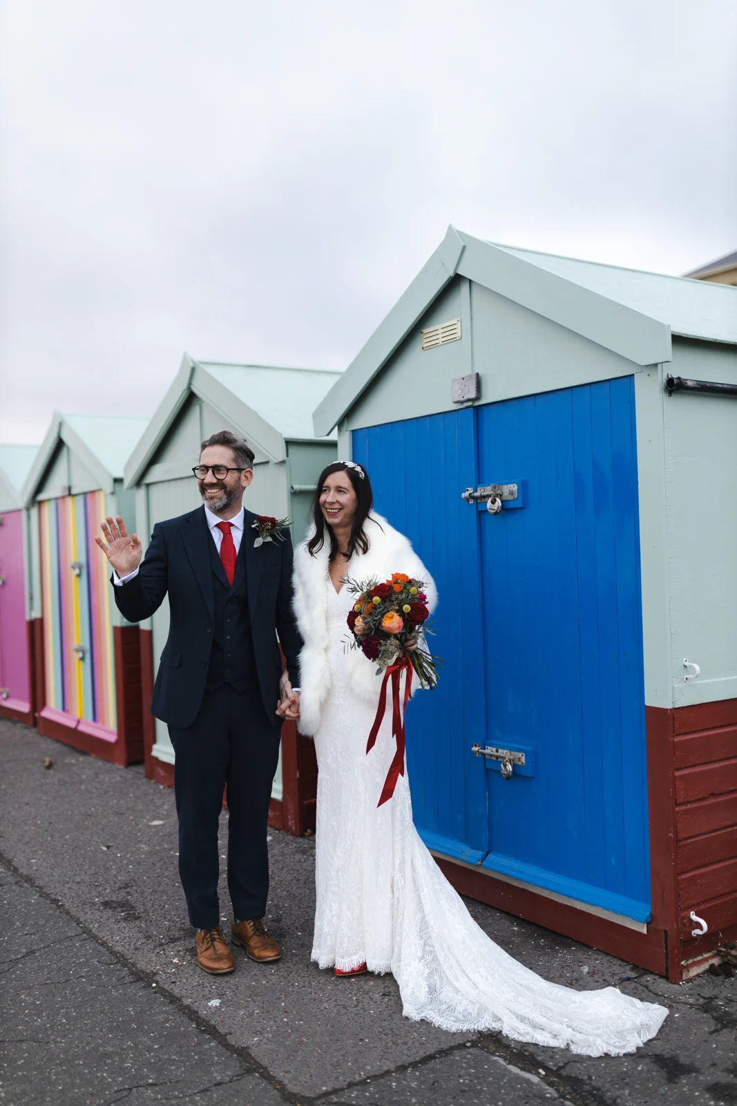 A wedding couple walking beside colourful beach huts in Hove.