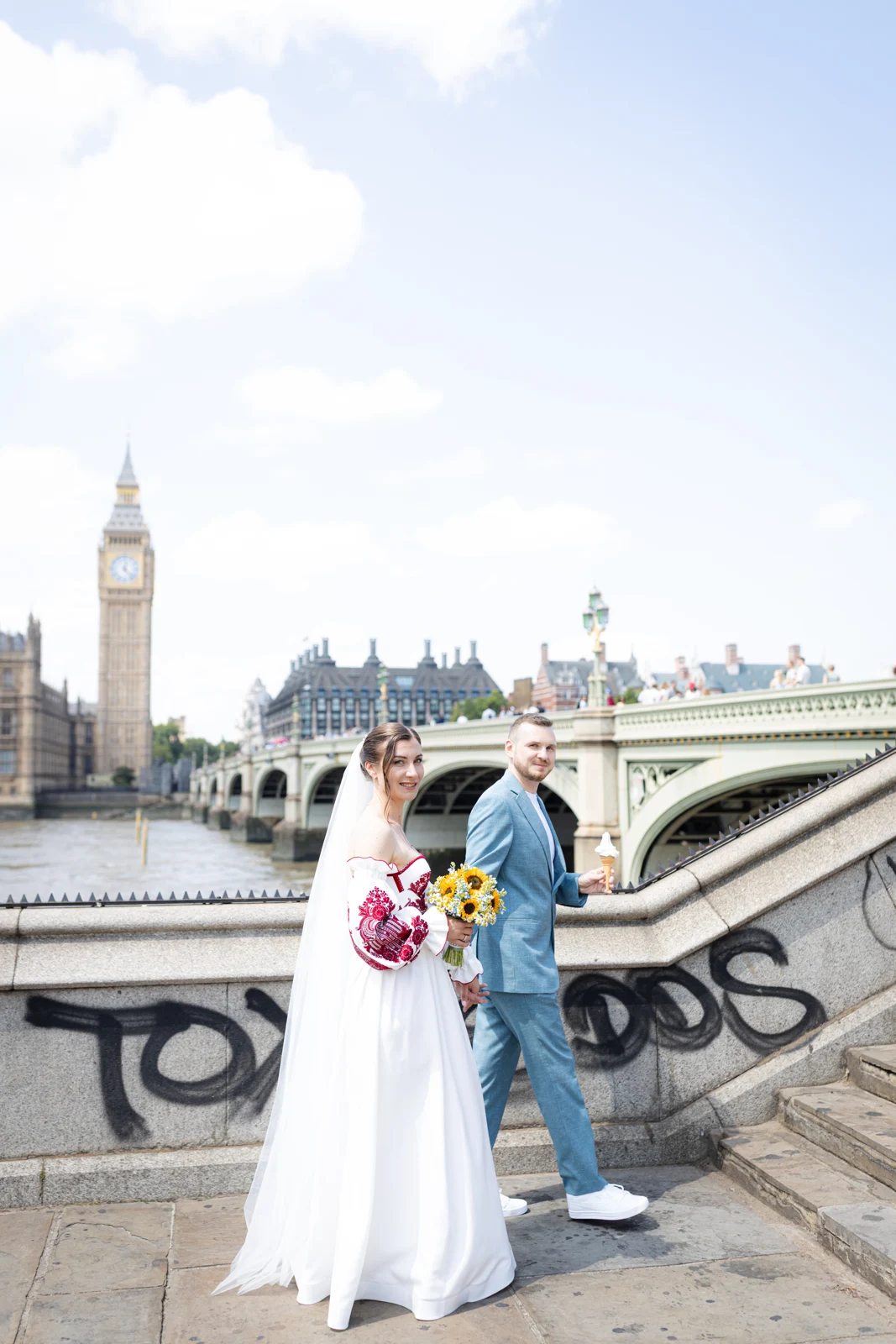 Lena and Arturs holding hands on the steps beside Westminster Bridge, with Big Ben behind them.