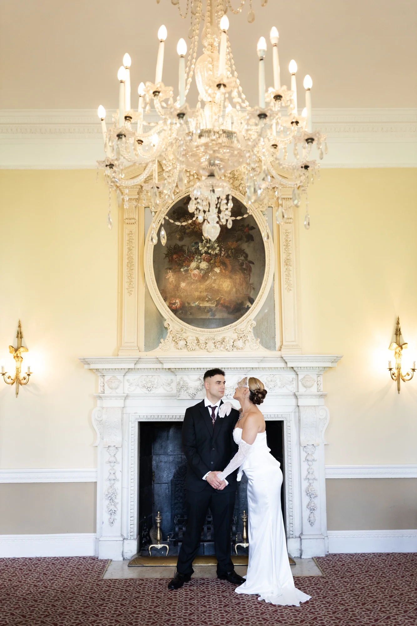 A bride and groom holding hands beneath a chandelier in front of an ornate fireplace.