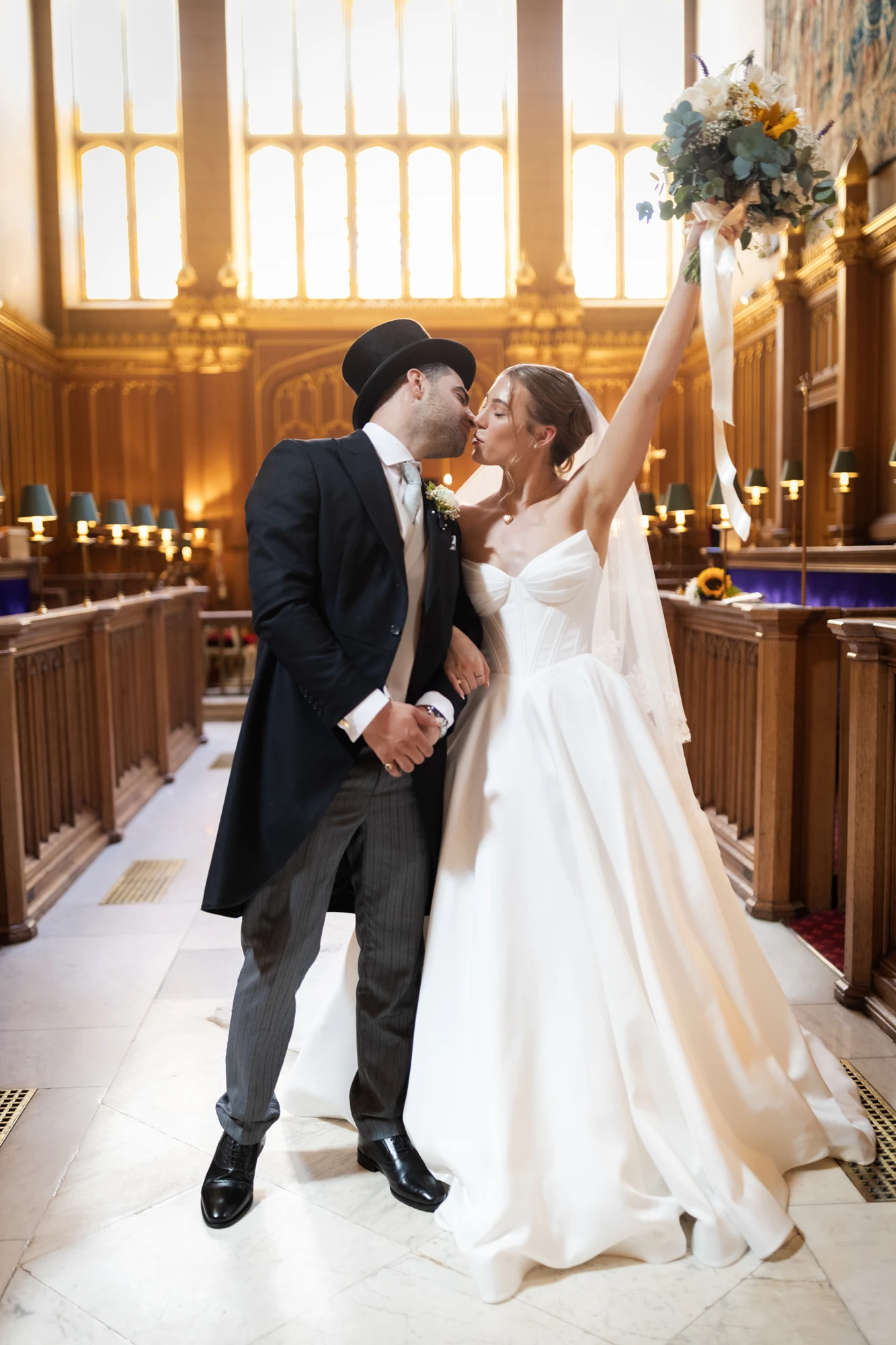 Newlyweds kissing in the chapel aisle as the bride raises her bouquet.