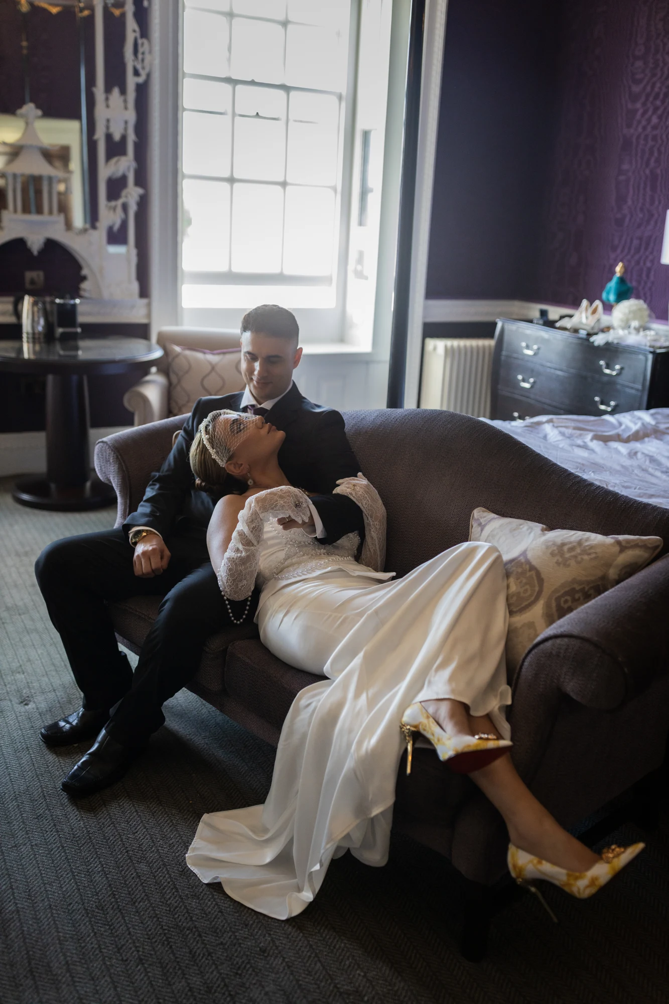 A bride and groom relaxing together on a sofa beside a window.